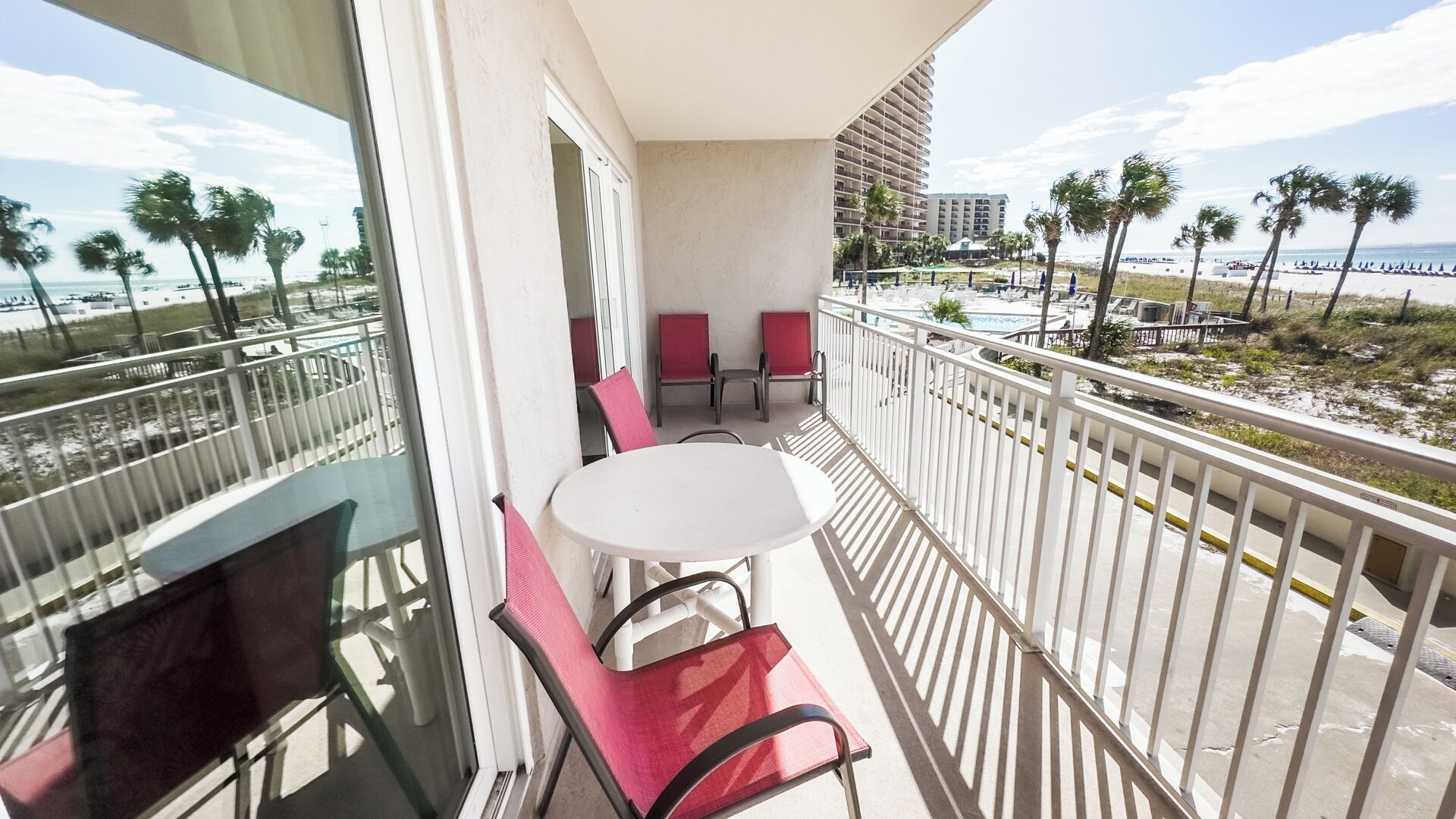 Un balcon avec une table et des chaises, donnant sur une plage bordée de palmiers et sur un bâtiment d'hôtel en arrière-plan.