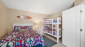 Un dormitorio con una cama litera, una cama con colcha estampada, una mesita de noche con lámpara y una puerta.