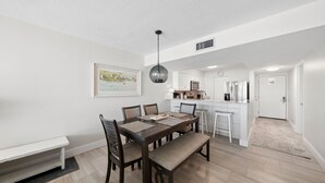 A dining area with a wooden table and chairs, a bench, a pendant light, a kitchen with white cabinets, and a door leading to another room. - C602 Sunset View, 3 Pools, Gulf Front (Panama City)