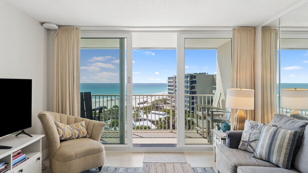 A balcony with a view of the beach and ocean, a television, a sofa, and a lamp. - C602 Sunset View, 3 Pools, Gulf Front (Panama City)