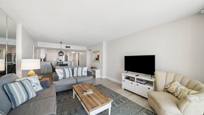 A modern living room with a grey sofa, a beige sofa, a coffee table, a TV on a stand, and a kitchen in the background. - C602 Sunset View, 3 Pools, Gulf Front (Panama City)