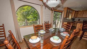 Un coin repas avec une table en bois dressée pour six personnes, entourée de chaises en bois. Une grande fenêtre offre une vue sur la verdure environnante.