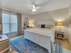 Un dormitorio con cama, mesitas de noche, una lámpara de noche, ventilador de techo, ventana con cortinas y alfombra estampada.