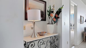 A hallway with a white console table, a lamp, a framed picture, and a potted plant with flowers.