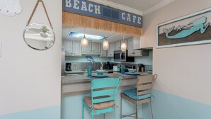 A kitchen with a 'BEACH CAFE' sign, a microwave, and two teal chairs.