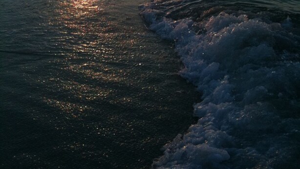A beach scene at sunset with waves lapping onto the shore, a pier extending into the water, and buildings in the background.