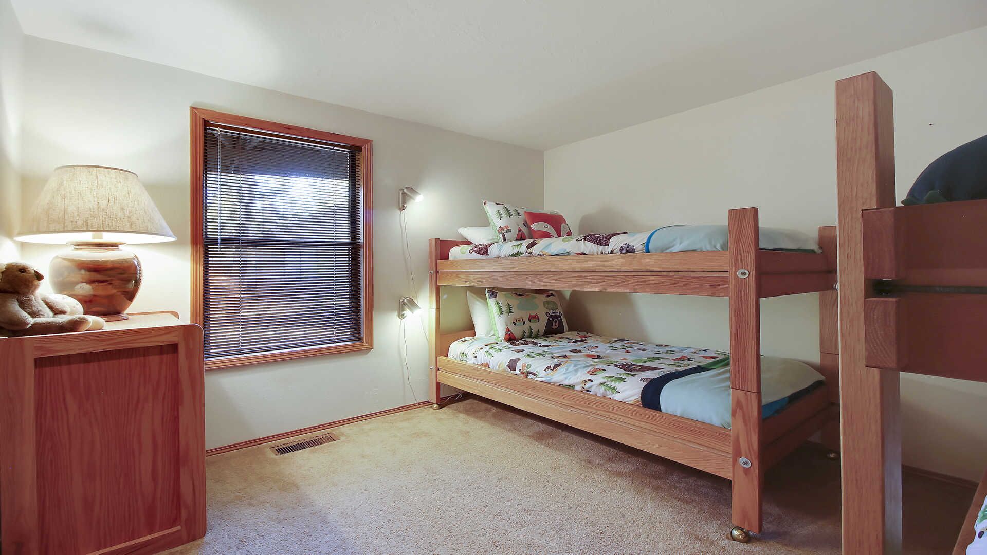 A bunk bed room with a lamp, a teddy bear, and a window with blinds.
