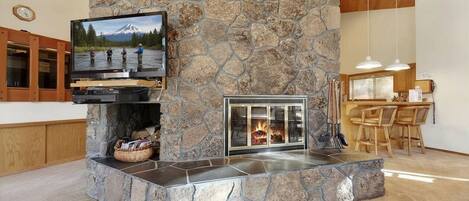 A stone fireplace with a TV above it, a stone wall, a kitchen area with wooden cabinets, and a dining table with chairs.