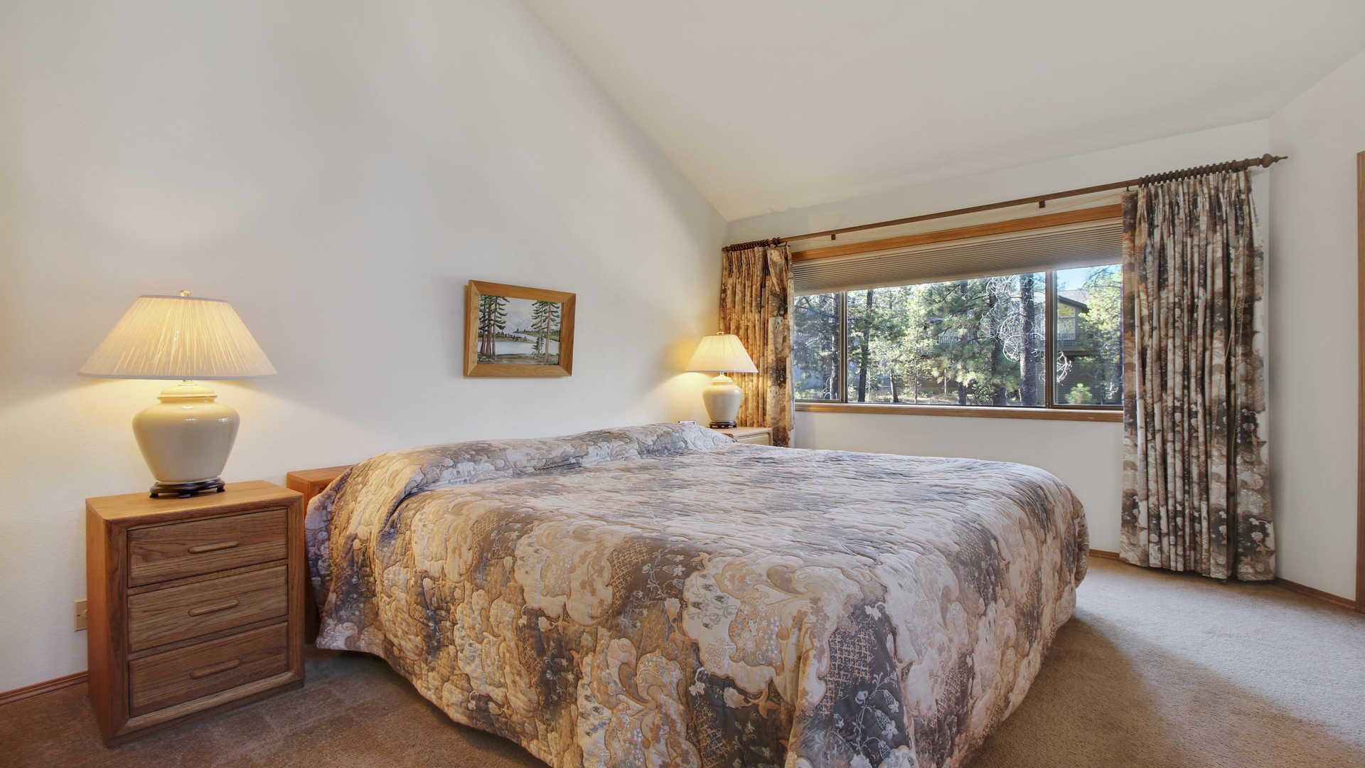 A bedroom with a large bed, two bedside tables with lamps, a window with curtains, and a framed picture on the wall.