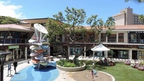Un'area esterna con un complesso giochi d'acqua, zone relax e un edificio con negozi sullo sfondo.
