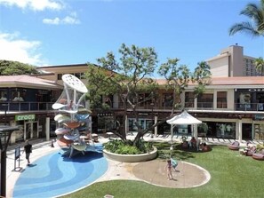 Uma área externa com uma estrutura de recreação aquática, áreas de estar e um edifício com lojas ao fundo.
