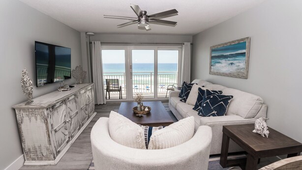 A living room with a view of the ocean, a ceiling fan, a flat-screen TV, a white sofa with patterned pillows, and a wooden coffee table. - D506 Corner unit, 2 balconies, Fantastic view of Pool and Gulf (Panama City Beach)