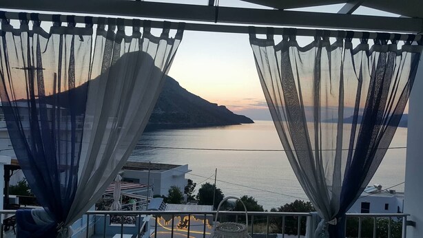 Un balcón con cortinas ligeras, vista a un mar tranquilo y una montaña en la lejanía.