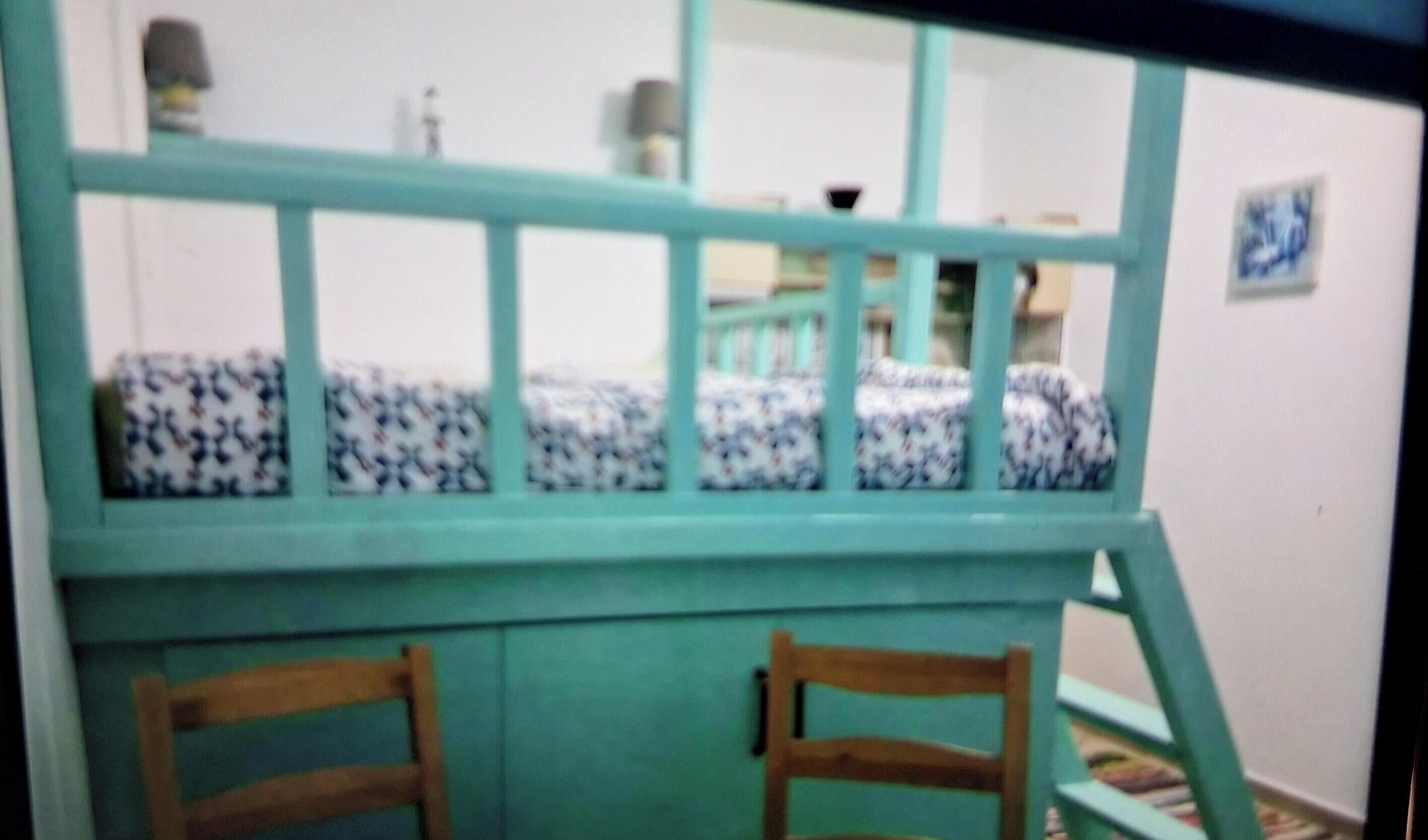 A bunk bed with a patterned mattress, a teal frame, and wooden chairs in a room with a white wall and a small picture.
