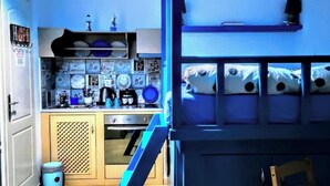 Cocina compacta con fregadero, estufa y diversos utensilios de cocina. Se observa un cesto de basura pequeño y una estantería con objetos decorativos.