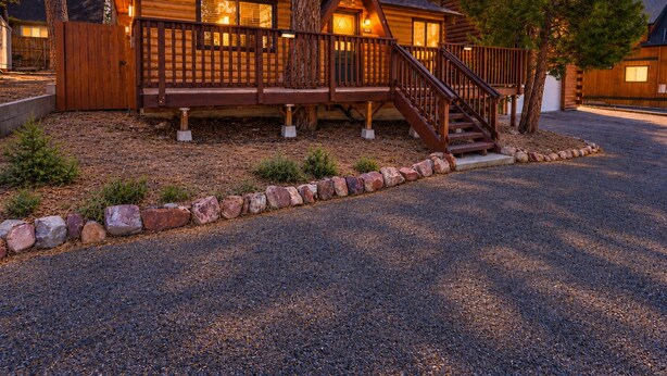 A wooden cabin with a deck and stairs leading to the entrance, surrounded by trees and a gravel driveway.