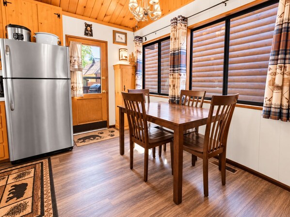 A wooden interior with a stainless steel refrigerator, a dining table with chairs, and a window with curtains.