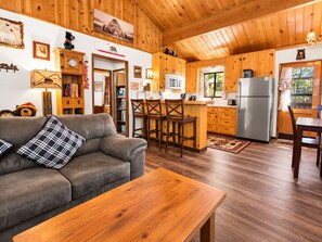 A cozy cabin interior with a wooden ceiling, a sofa with a plaid pillow, a wooden table, a kitchen area with a refrigerator, and a dining table.