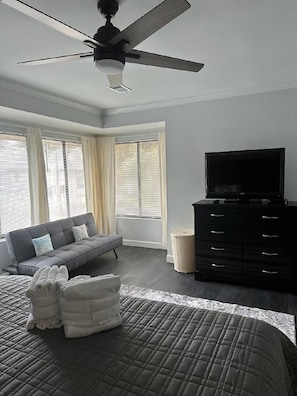 Ein Schlafzimmer mit einem Bett, einem grauen Sofa, einem Fernseher auf einer schwarzen Kommode und einem Deckenventilator.