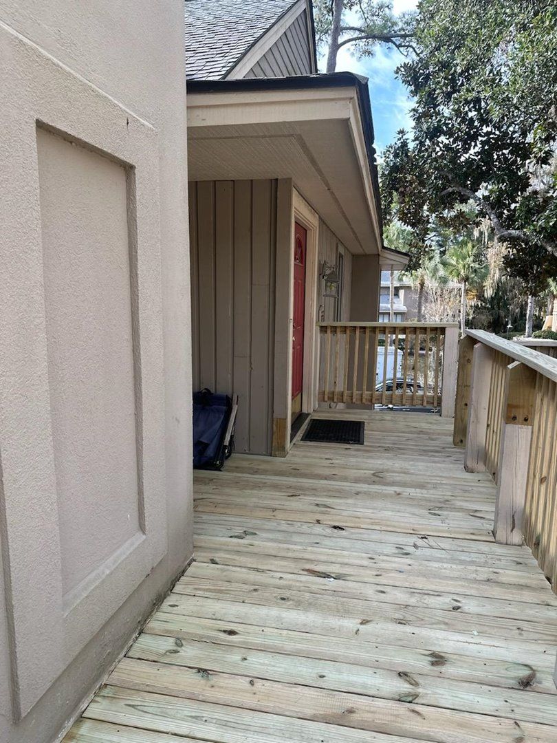 A wooden deck leading to a building with a red door, a small porch, and a view of trees and a parked car.
