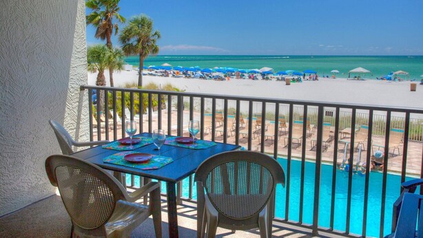 Un balcón con una mesa dispuesta para dos, con vista a una playa con sombrillas y una piscina.
