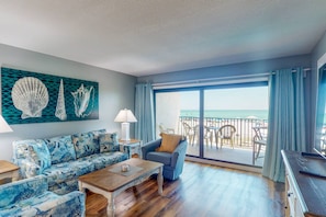 Un soggiorno con divano, poltrona e tavolino, un quadro raffigurante conchiglie e un balcone con vista sulla spiaggia.
