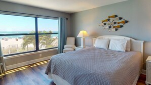 Una habitación de hotel con un ventanal grande, una cama, una mesita de noche, una lámpara y un adorno en la pared.