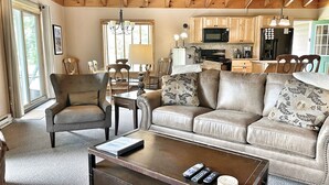 A cozy living room with a sofa, armchair, and coffee table, wooden ceiling beams, and a kitchen in the background. - Mountain Home, Views, Near Whiteface, Lake Placid (Wilmington)