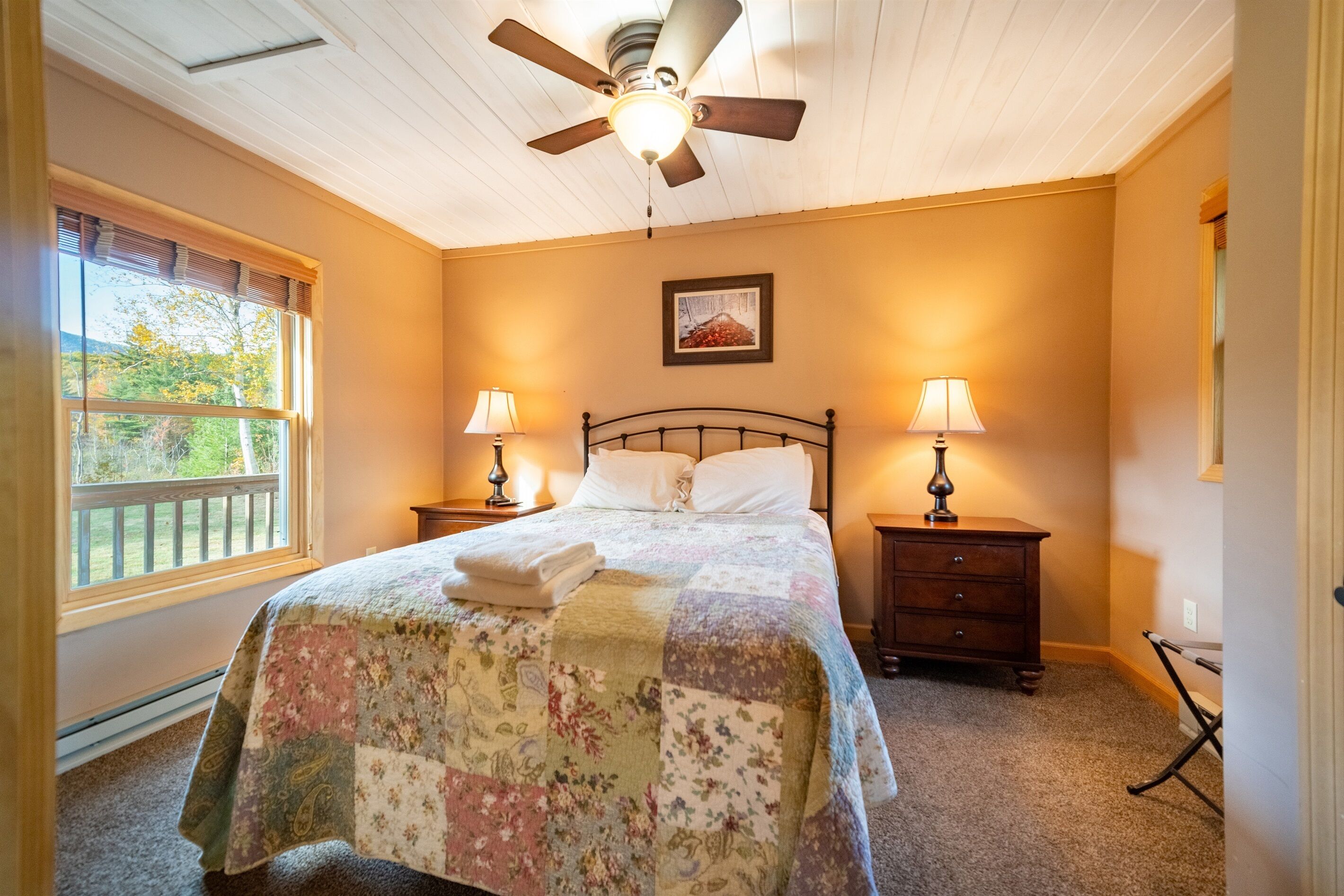 A bedroom with a bed, two bedside tables with lamps, a ceiling fan, a window with blinds, and a picture on the wall.