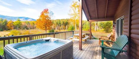 A wooden deck with a hot tub, picnic table, and chairs, overlooking a forested area with autumn foliage.