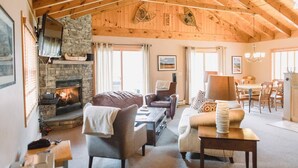 A cozy living room with a stone fireplace, wooden beams, and a dining area with a chandelier. - Mountain Home, Views, Near Whiteface, Lake Placid (Wilmington)