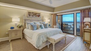 Una camera da letto con un letto grande, una ventola a soffitto, un comodino con una lampada, una panca e vista sull'oceano tramite una porta scorrevole in vetro.