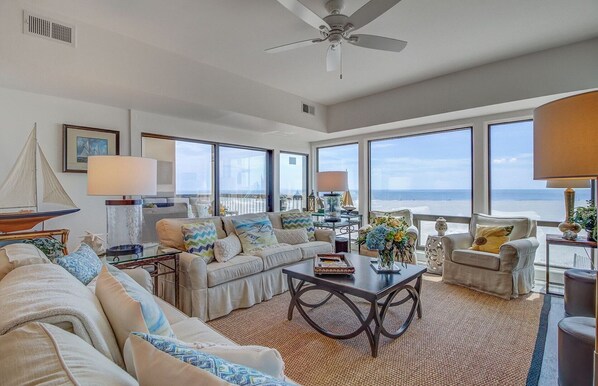 A spacious living room with a large window overlooking the beach, a ceiling fan, and a view of the ocean.