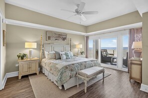 Une chambre à coucher comprenant un lit, des tables de chevet, un banc, un ventilateur de plafond et une vue sur la plage, accessible par une porte coulissante.