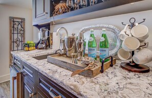 A kitchen with a marble countertop, a sink, and various kitchen appliances. There are bottles of water and a tray with cups on the counter.