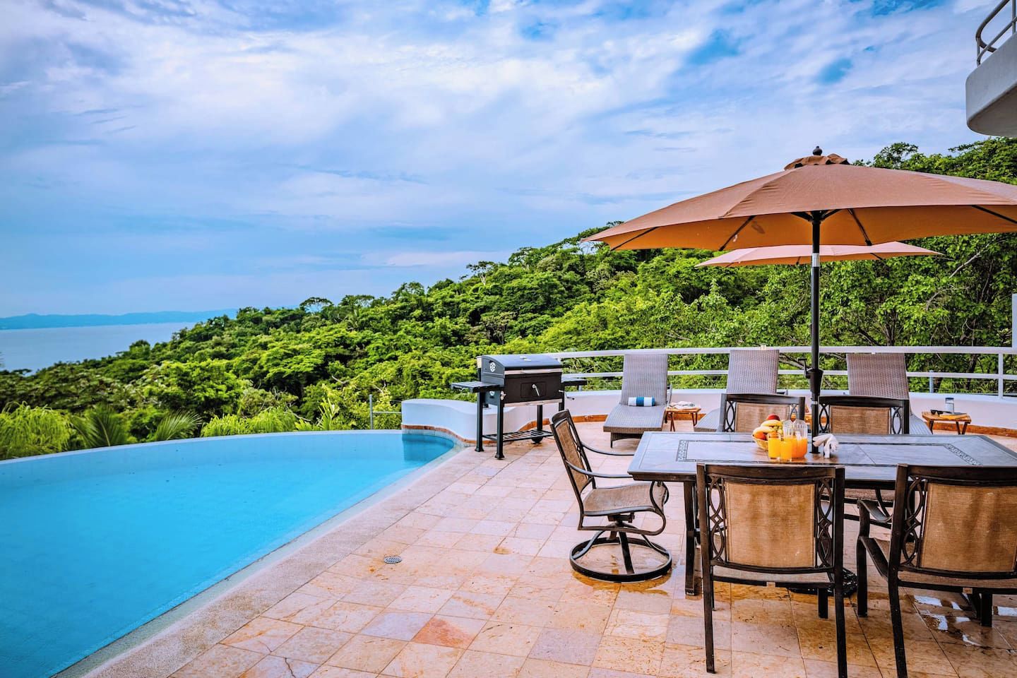 A poolside area with a view of the sea, outdoor furniture, and a grill.
