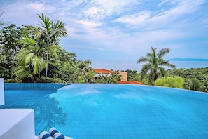 A clear blue infinity pool with a view of the sea and mountains, surrounded by lush greenery and a building with a red roof.