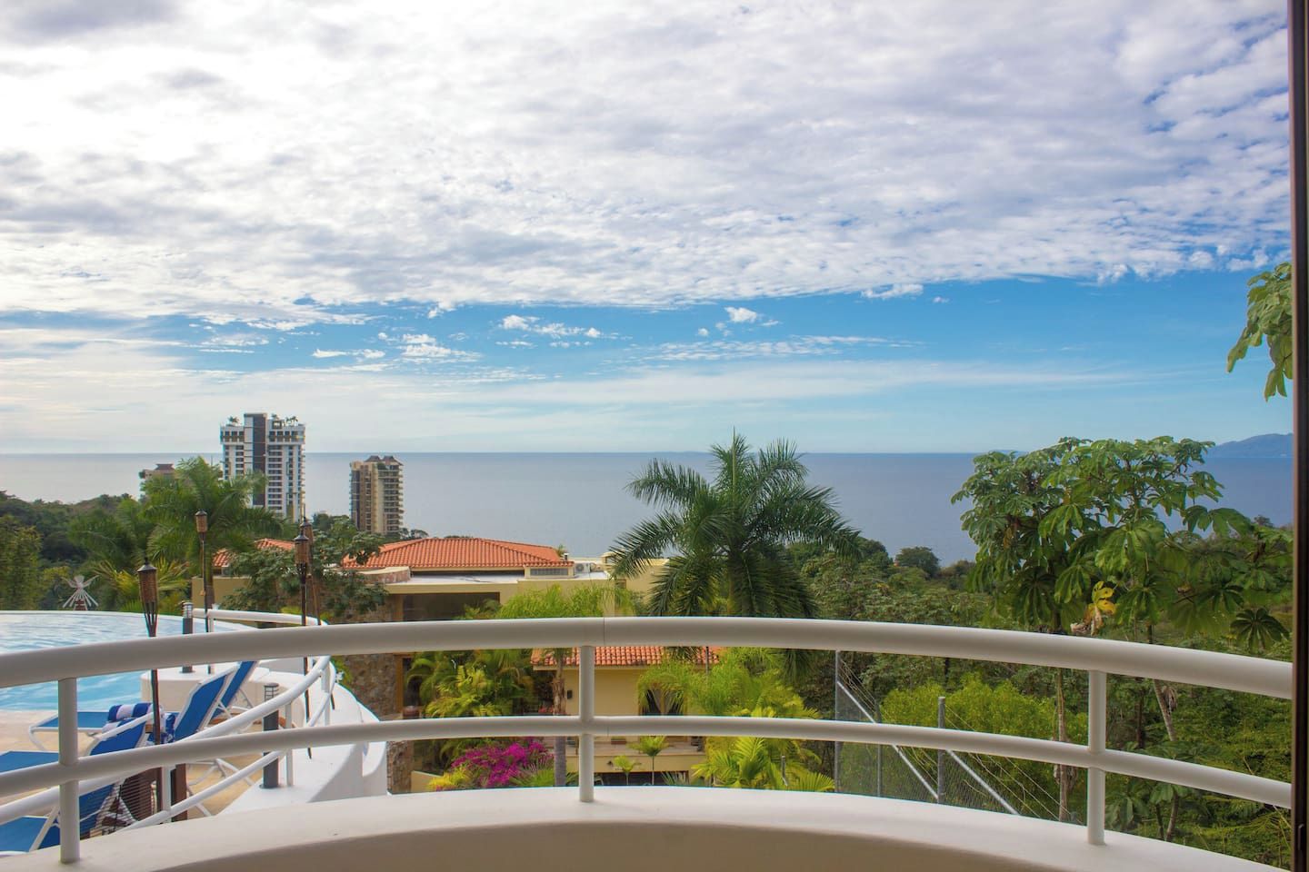 A balcony with a view of a swimming pool, palm trees, and distant buildings.