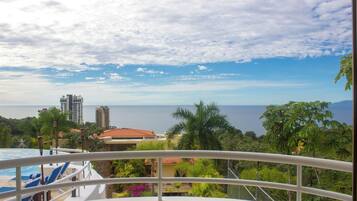 A balcony with a view of a swimming pool, palm trees, and distant buildings.