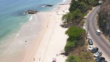 A beach with a road alongside, cars parked on the side, and people walking on the beach.
