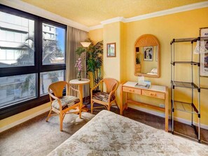 Hotelkamer met een bed, bureau, stoel, lamp, plant en raam met uitzicht op palmbomen.