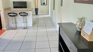 A hallway with a kitchen area, a dining area with stools, a TV mounted on the wall, and a painting of a pier. - Newly Remodeled Home - Steps away from white sand beaches!!! (Lauderdale-By-The-Sea)