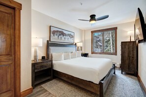 A bedroom with a large bed, two bedside tables, a ceiling fan, a window with a view, and a wooden wardrobe.