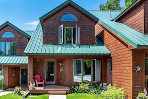 A two-story wooden building with a green roof, a front porch with a red chair, and a flag.
