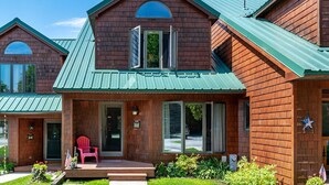 A two-story wooden house with a green metal roof, a front porch with a red chair, and a mailbox.
