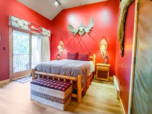 Quarto com uma cama, um banco, duas mesas de cabeceira com abajures, uma parede vermelha e uma janela com cortinas.