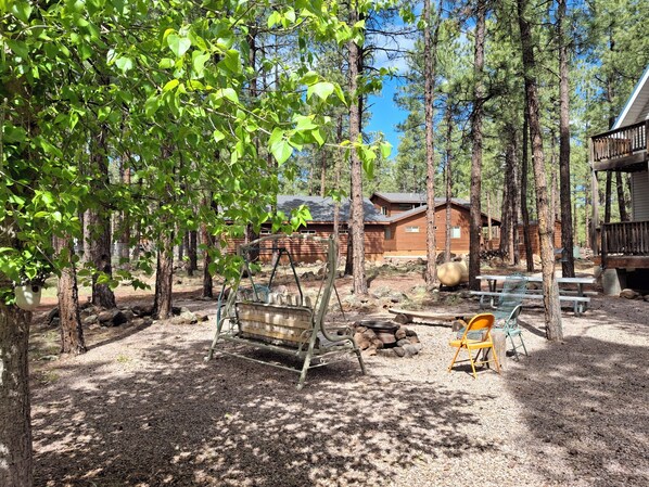 A rustic cabin surrounded by tall trees, a swing set, a fire pit, and outdoor seating.