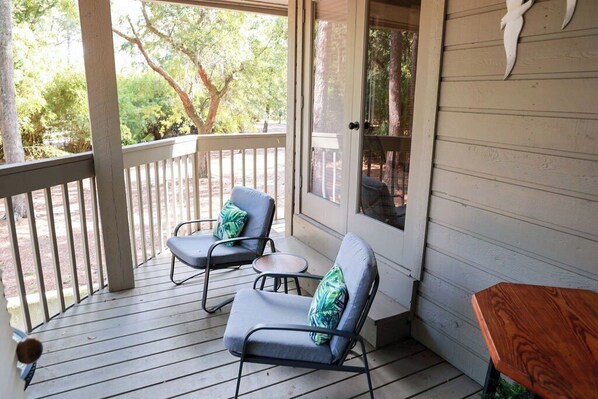 A covered porch with two lounge chairs and a small table, overlooking a wooded area.