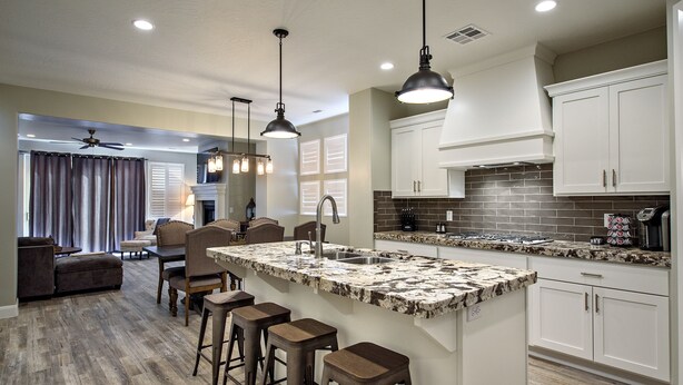 A modern kitchen with white cabinets, a large island with bar stools, pendant lighting, and a view into the living area. - Stunning Red Rock Views - 4BR Poolside Fire Pit (St. George)