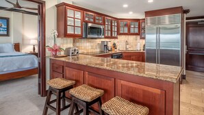 Una cocina con gabinetes de madera, un refrigerador de acero inoxidable, encimeras de granito y tres taburetes de mimbre.